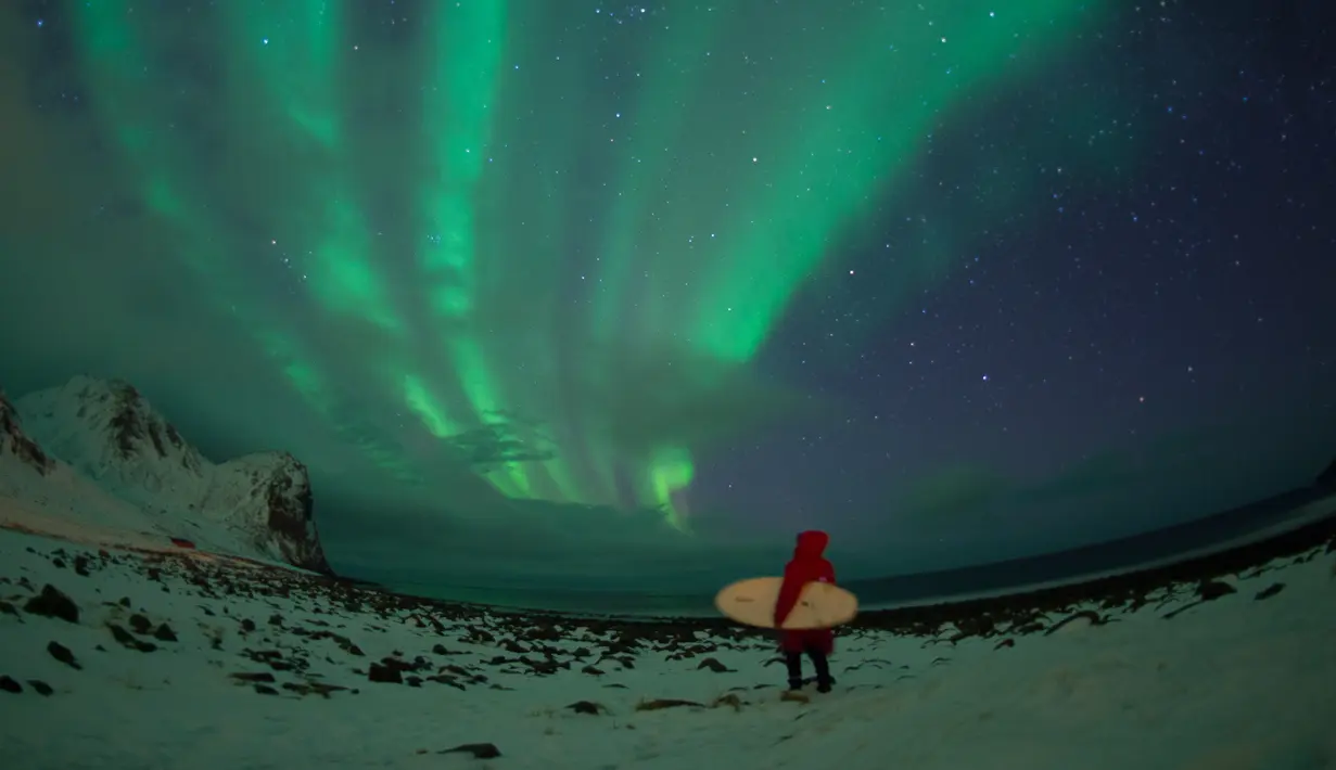 Peselancar berdiri dibawah cahaya Aurora Borealis di pantai bersalju Flackstad di Kepulauan Lofoten, Lingkar Arktik, pada 10 Maret 2016. Peselancar dari seluruh dunia datang ke Lofoten untuk berselancar di kondisi ekstrim. (AFP/Olivier Morin)