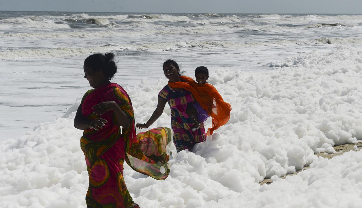 Penduduk setempat berjalan di atas limbah busa yang disebabkan polutan saat bercampur dengan ombak di pantai di Chennai (29/11/2019). Limbah busa tersebut  membuat penduduk dan pengunjung pantai merasa sakit. (AFP/Arun Sankar)