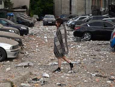 Seorang penduduk lokal berjalan di antara mobil-mobil yang rusak saat meninggalkan bangunan tempat tinggal bertingkat, yang sebagian hancur setelah serangan pesawat tak berawak pada malam hari di Kyiv pada 30 Mei 2023. (Sergei SUPINSKY/AFP)