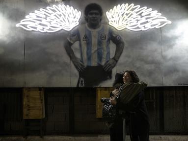 Perempuan berpelukan di dekat mural mendiang legenda sepak bola dunia, Diego Maradona di Buenos Aires, Argentina, Selasa malam (23/11/2021). Peringatan pertama kematian Maradona digelar pada 25 November 2021. (AP Photo/Rodrigo Abd)