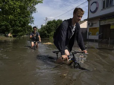 Penduduk setempat mencoba mengendarai sepeda mereka di sepanjang jalan yang banjir setelah bendungan Kakhovka meledak semalaman, di Kherson, Ukraina, Selasa, 6 Juni 2023.(AP Photo/Evgeniy Maloletka)