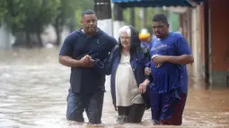 Dua pria membantu seorang wanita menyeberangi jalan yang banjir di Sao Paulo, Brasil, Senin, (10/2/2020). Hujan deras yang membanjiri kota, menyebabkan pinggir sungai utama meluap. (AP Photo/Andre Penner)