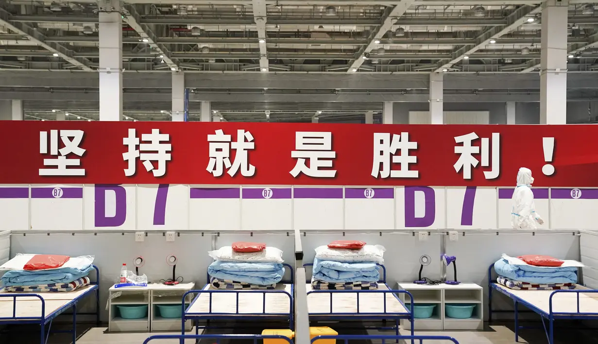 Seorang pekerja dengan pakaian pelindung berjalan melewati spanduk bertuliskan "Ketekunan adalah kemenangan!" di rumah sakit darurat dan fasilitas karantina di Pusat Pameran dan Konvensi Nasional di Shanghai (11/4/2022). (AP Photo)