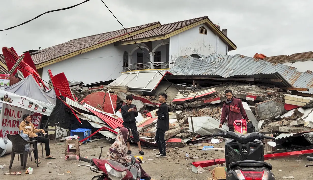 Penduduk berdiri di dekat bangunan yang diratakan oleh gempa bumi di Mamuju, Sulawesi Barat, Jumat (15/1/2021). Korban meninggal dikhawatirkan akan terus bertambah, karena beberapa laporan menyebutkan kemungkinan ada korban yang masih terjebak di bawah reruntuhan. (AP Photo/Rehan)