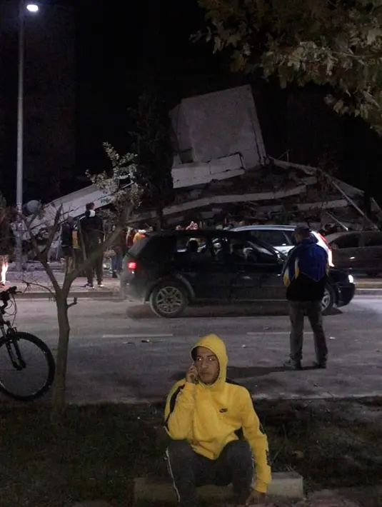Seorang pria berbicara di ponsel ketika yang lain melihat kerusakan bangunan setelah gempa bumi di Durres,  Albania barat, Selasa (26/11/2019). Gempa bumi bermagnitudo 6,4 mengguncang Albania, Selasa dini hari yang menyebabkan beberapa bangunan dan gedung permukiman runtuh. (AP Photo)