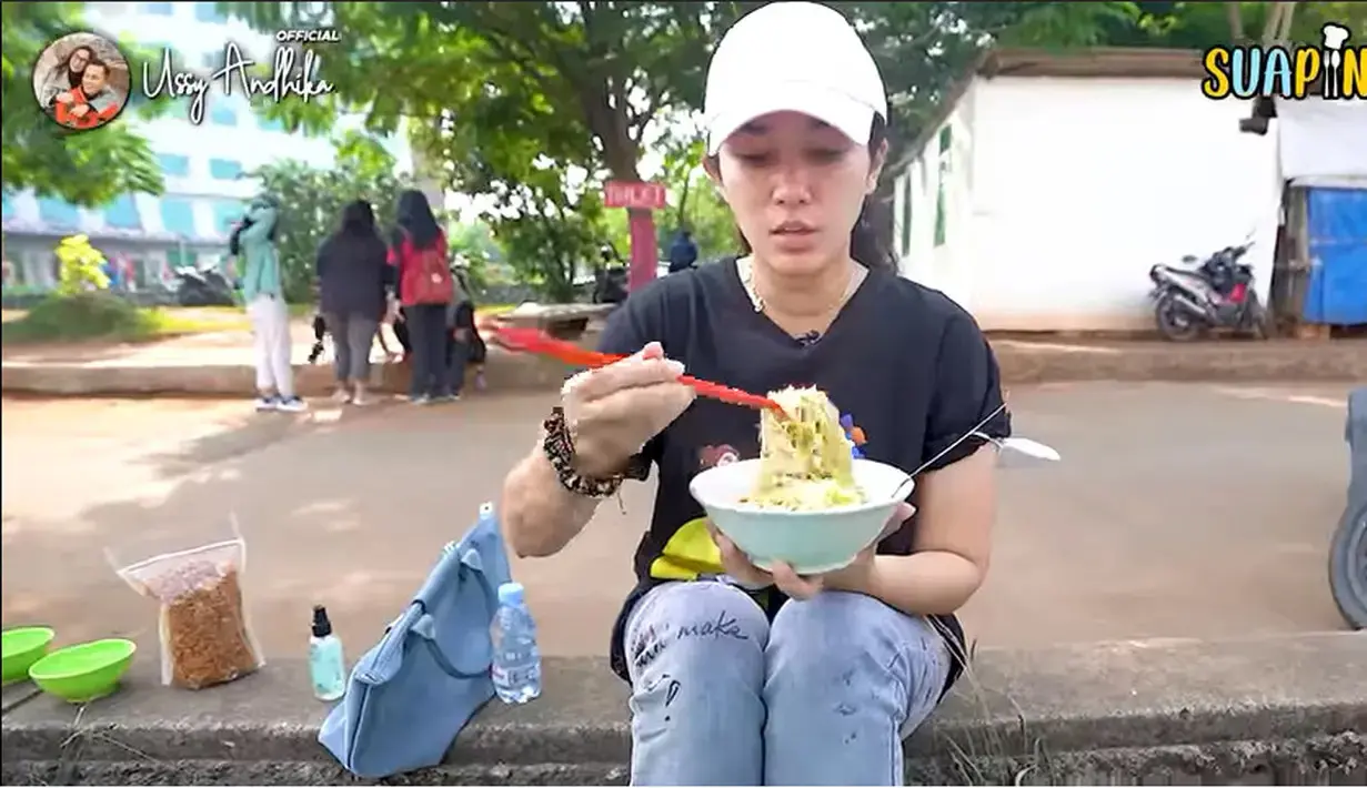 Menikmati mie ayam pinggir jalan dengan gaya santai ngemper. Ussy tampak santai duduk di beton pembatas jalan dan menikmati makanan pedagang kaki lima. [Youtube/Ussy Andhika Official]