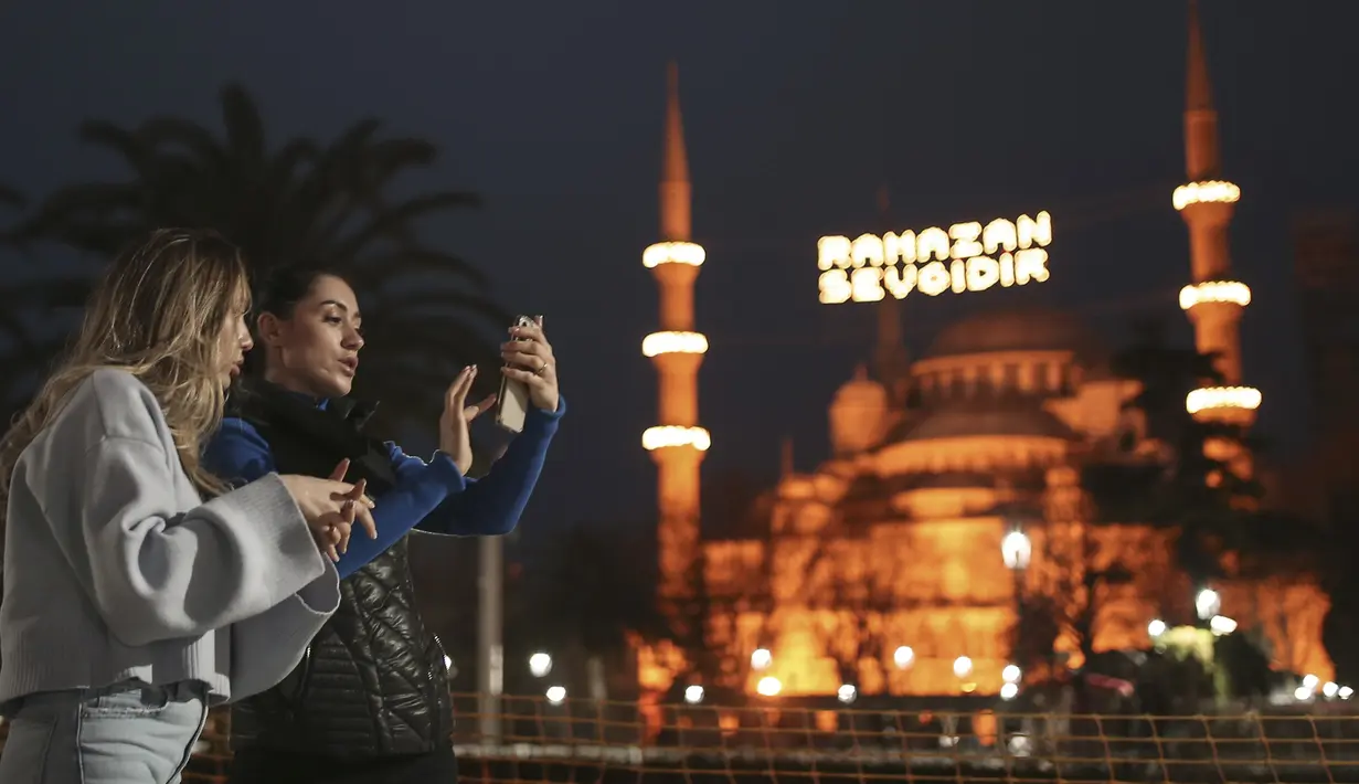 Dua wanita berdiri di dekat Masjid Sultan Ahmed yang ikonik, lebih dikenal sebagai Masjid Biru, dihiasi dengan lampu dan slogan bertuliskan "Ramadhan adalah cinta," menandai bulan Ramadhan, di distrik bersejarah Sultan Ahmed di Istanbul (13/4/2021). (AP Photo/Emrah Gurel)
