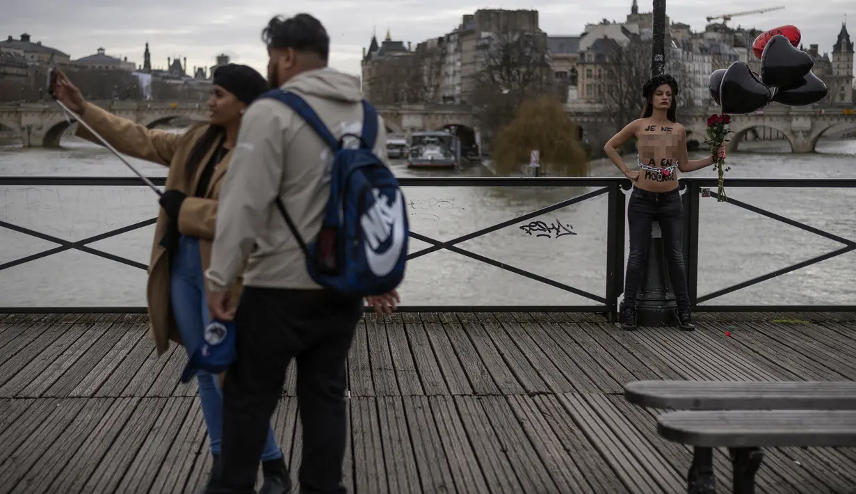 Pasangan berselfie dengan latar anggota gerakan feminis Femen yang dirantai selama aksi untuk mengecam kekerasan terhadap perempuan pada hari Valentine di pont des Arts, Paris (14/2/2020). (AFP Photo/Lionel Bonaventure)