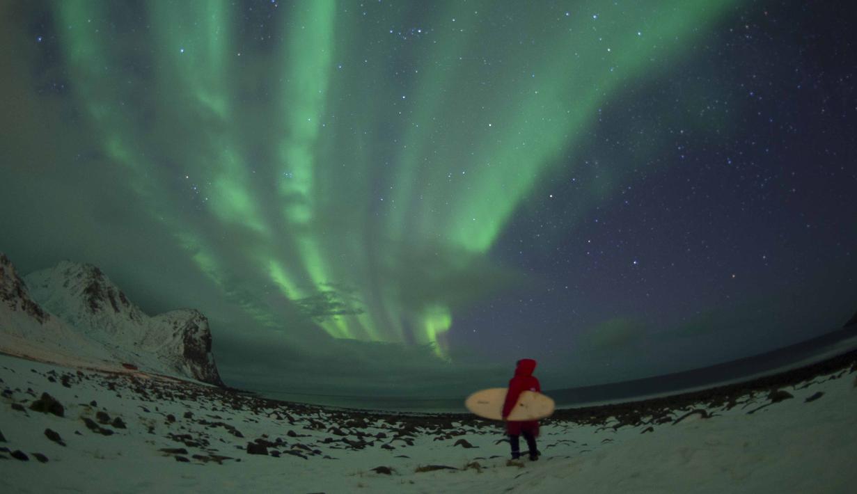 Seorang peselancar berdiri menatap sinar aurora sebelum beraksi di pantai Unstad, Lofoten, Arctic Circle, (10/3/2016). (AFP/Olivier Morin)