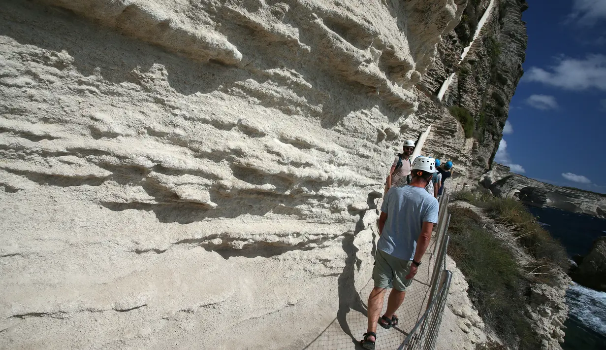 Turis menyusuri jalan setapak di kaki tebing sebelum menaiki tangga Raja Aragon di Bonifacio di pulau Corsica, Mediterania Prancis (19/9/2021). Tangga Raja Aragon digali dengan tangan di tebing batu kapur di suatu malam oleh pasukan Raja Aragon tahun 1420. (AFP/Pascal Pochard-Casabianca)