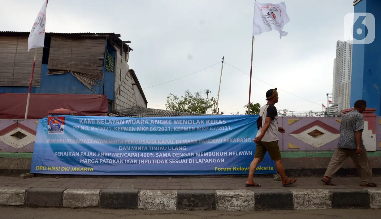 Warga melintasi spanduk penolakan PP Nomor 85 Tahun 2021 KKP terkait tarif atas jenis PNBP di Pelabuhan Muara Angke, Jakarta, Sabtu (9/10/2021). Dengan terbitnya PP Nomor 85 Tahun 2021, PNBP untuk para nelayan mengalami kenaikan sebesar 600 persen dari tarif biasanya. (merdeka.com/Imam Buhori)