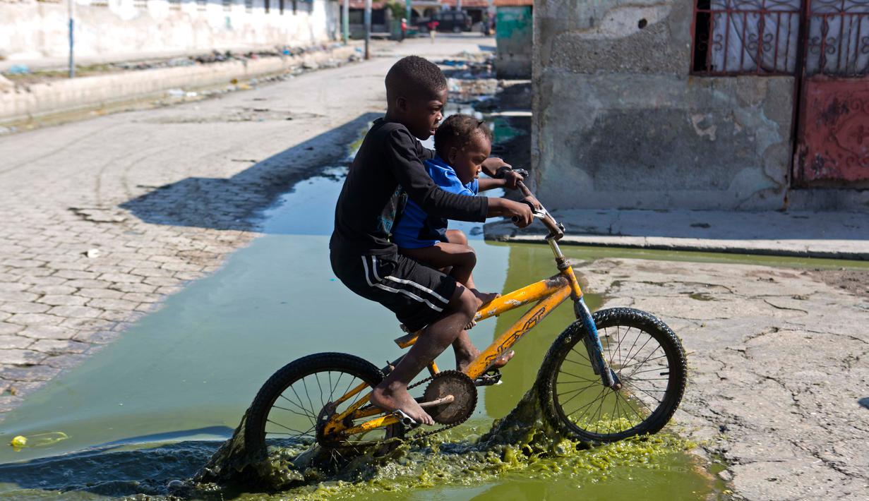 Dua anak melewati genangan air limbah saat naik sepeda melewati daerah Cite Soleil di Port-au-Prince, Haiti (17/3). Cite Soleil awalnya dikembangkan sebagai kota kumuh dan tumbuh hingga sekitar 200.000 hingga 400.000 penduduk. (AP Photo/Dieu Nalio Chery)