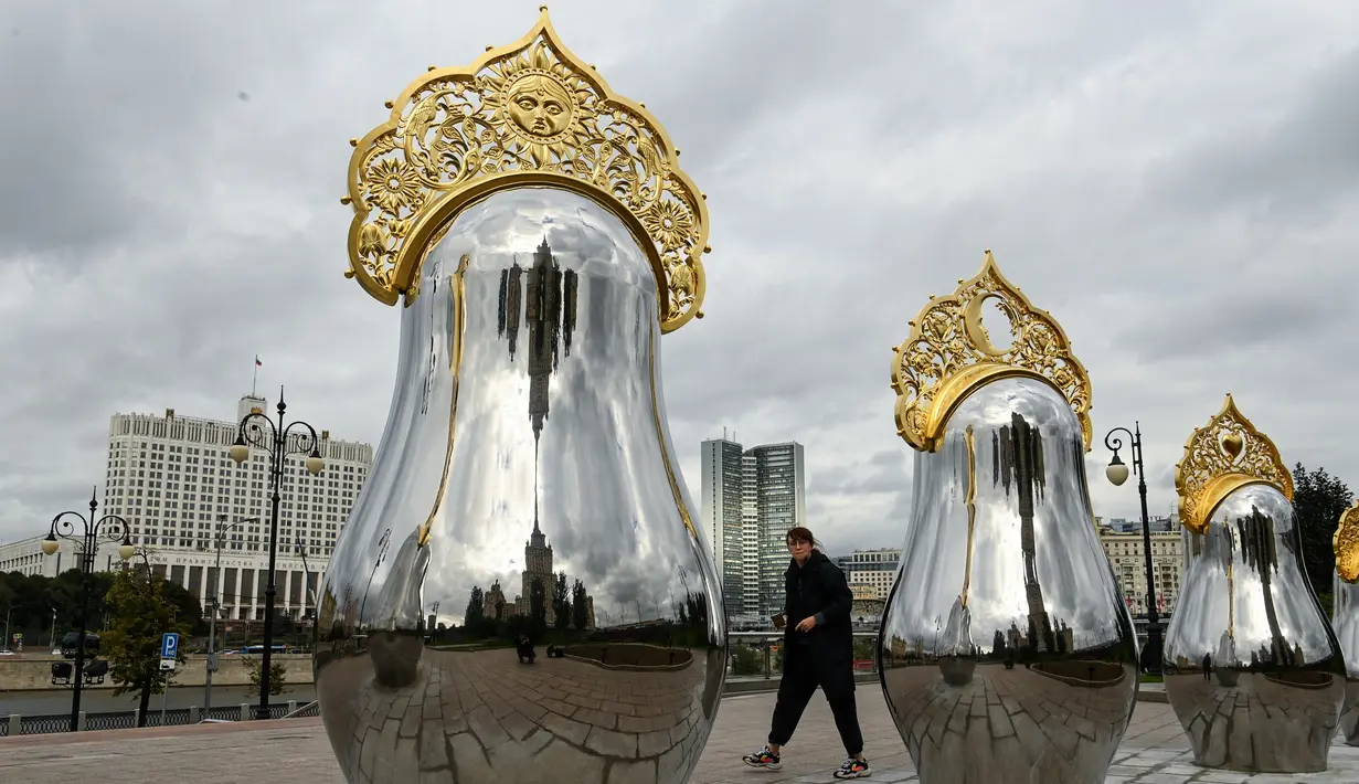 Seorang wanita berjalan melewati "benda seni Matryoshki", tujuh boneka bersarang tradisional Rusia "matryoshka" yang dicerminkan dengan hiasan kepala "kokoshnik", dengan gedung Pemerintah Rusia terlihat di latar belakang, di pusat kota Moskow (9/9/2021).  (AFP/Kirill Kudryavtsev)