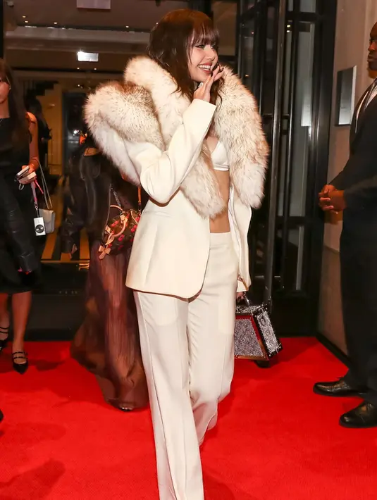 Setelah tampil dramatis dengan gaun couture di red carpet, Lisa beralih ke power dressing yang lebih playful. Ia terlihat meninggalkan hotel dengan setelan cream suit berpotongan tailored dengan low-rise flared pants. Di balik blazer, terselip satin bralette yang memberi sentuhan sensual. Aksesori seperti platform heels dari Stuart Weitzman, trunk bag berkilau, dan perhiasan berlian memperkuat aura glam yang effortless. [@voguehongkong].