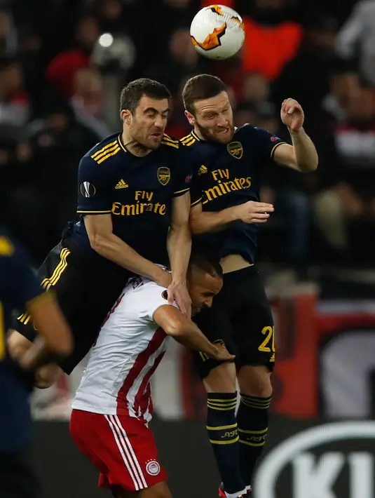 Bek Arsenal, Sokratis Papastathopoulos dan rekannya, Shkodran Mustafi melompat saat berebut bola dengan pemain Youssef El-Arabi  pada leg pertama babak 32 besar Liga Europa di Georgios Karaiskakis Stadium, Kamis (20/2/2020). Arsenal menaklukkan Olympiakos Piraeus 1-0. (AP/Thanassis Stavrakis)