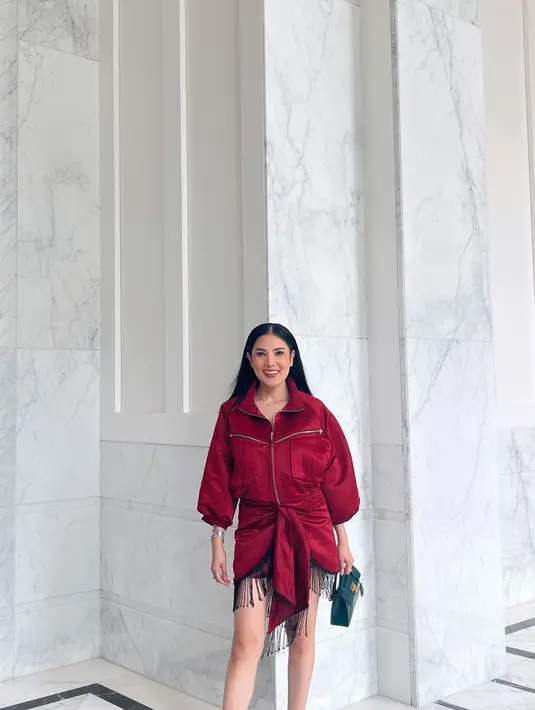 Nindy Ayunda tampil elegan dan stylish mengenakan wrap dress merah dengan aksen fringe di bagian bawahnya. Dress-nya kemudian dipadukan dengan tas jinjing warna hijau yang kontras dengan penampilannya. [IG/@nindyayunda].