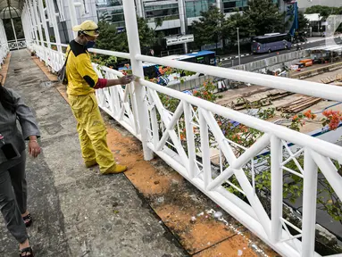 Warga melintasi pekerja Dinas Bina Marga DKI yang mengecat ulang tiang Jembatan Penyeberangan Orang (JPO) di kawasan Thamrin, Jakarta, Kamis (28/1/2021). Pengecatan ulang itu sebagai bagian dari perawatan JPO di Jakarta guna memberikan kesan bersih dan indah. (Liputan6.com/Faizal Fanani)