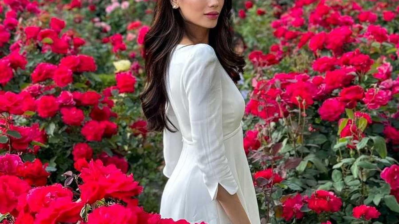 Sabrina mempesona dengan penampilan anggun dalam gaun putih, dikelilingi oleh kecantikan bunga mawar yang mekar di sekelilingnya.
