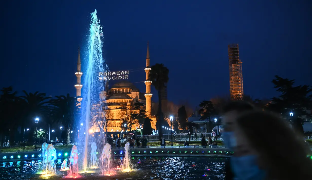 Sepasang suami istri berjalan melewati air mancur pada hari pertama bulan puasa Muslim Ramadhan di Sultanahmet Square dengan latar belakang Masjid Biru, di Istanbul (13/4/2021). (AFP Photo/Ozan Kose)