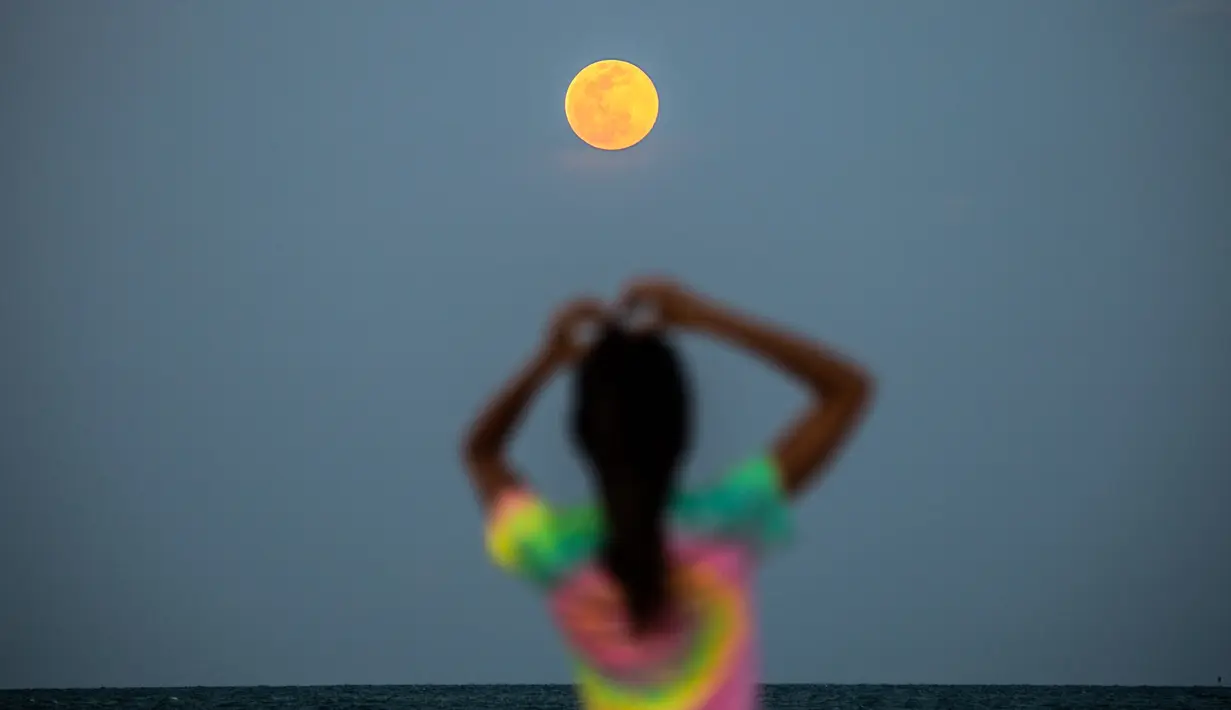 Bulan Purnama Super Pink terlihat di latar belakang saat orang-orang bersantai di pantai di Miami Beach, Florida, Senin (26/4/2021). Super Pink Moon merupakan fenomena alam ketika posisi bulan purnama berada terdekat dengan bumi. (CHANDAN KHANNA / AFP)