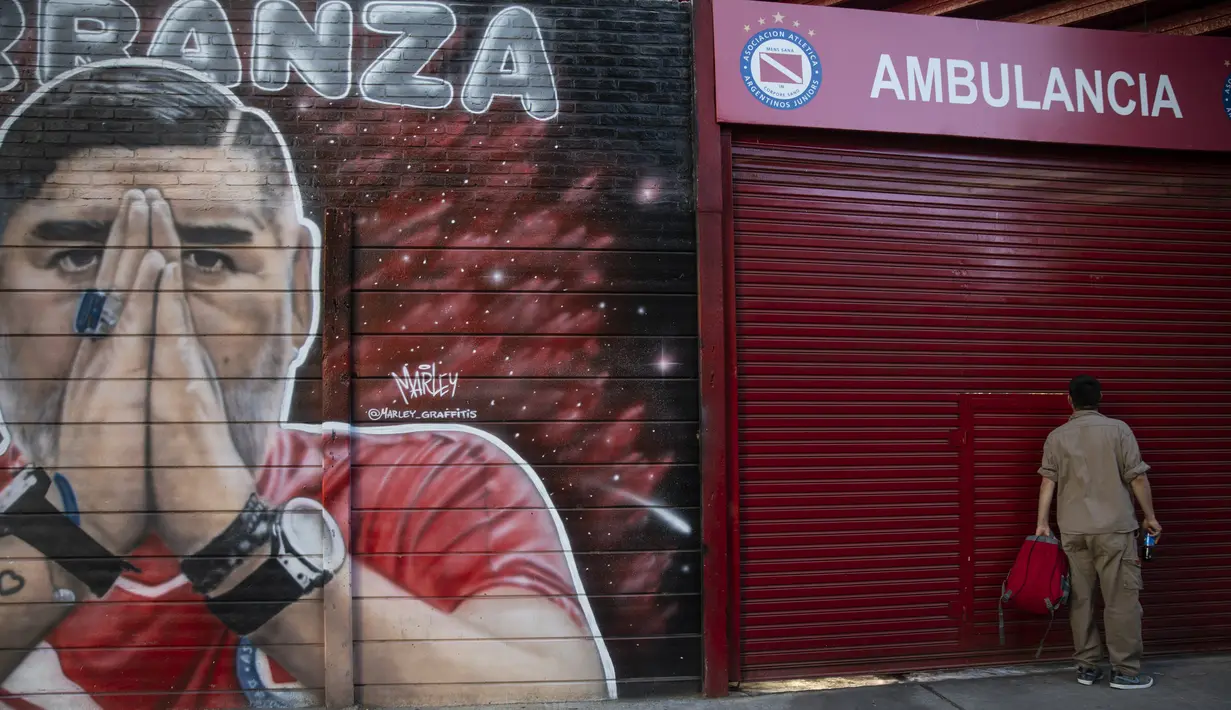 Seorang pekerja mengintip di dalam stadion Argentinos Juniors di sebelah mural mendiang legenda sepak bola dunia, Diego Maradona di Buenos Aires, Argentina, Selasa (23/11/2021). Peringatan pertama kematian Maradona digelar pada 25 November 2021. (AP Photo/Rodrigo Abd)
