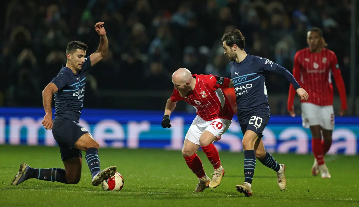 Gelandang tim divisi 4 Swindon Town, Jonny Williams (tengah) bersaing dengan pemain Manchester City Rodri (kiri) dan Bernardo Silva dalam duel putaran ketiga Piala FA 2021/22 di County Ground, Sabtu (8/1/2022) dini hari WIB. Manchester City menang telak 4-1 atas Swindon Town. (Adrian DENNIS / AFP)
