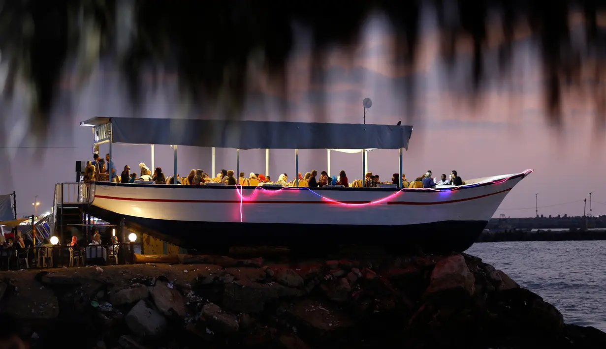 Warga Palestina menyantap hidangan sambil menikmati senja di sebuah restoran berbentuk perahu di tepi pantai Kota Gaza, Jalur Gaza, Rabu (2/8). Restoran tersebut berada di atas batu karang dengan hiasan lampu warna-warni. (MOHAMMED ABED / AFP)