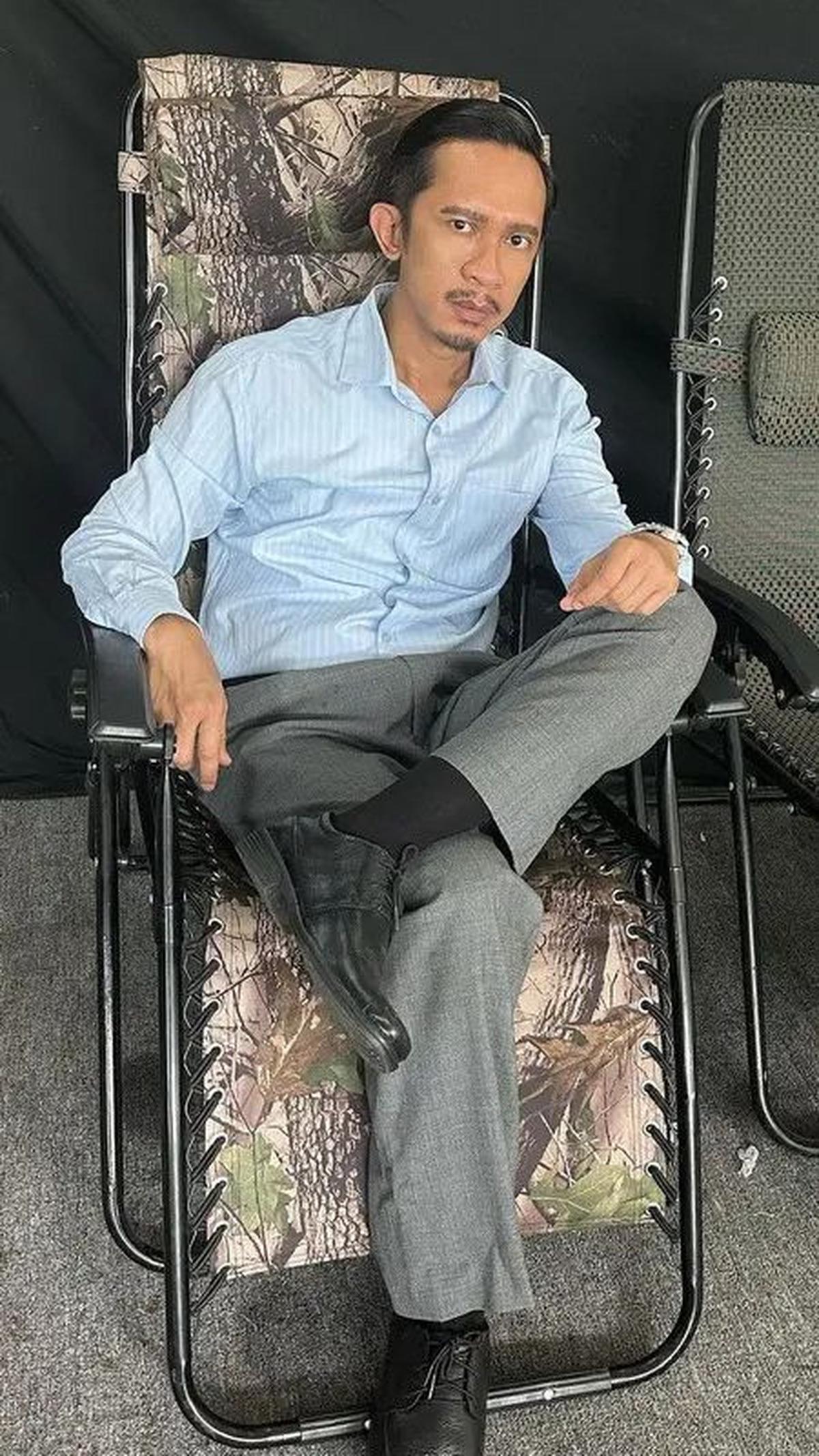 Aming tampil berbeda dengan kemeja biru muda, celana panjang abu-abu, dan alas kaki hitam.