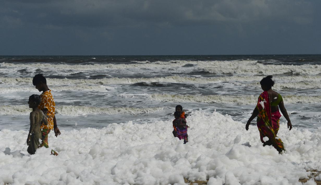 Penduduk setempat berjalan di atas limbah busa yang disebabkan polutan saat bercampur dengan ombak di pantai di Chennai (29/11/2019). Limbah busa tersebut  membuat penduduk dan pengunjung pantai merasa sakit. (AFP/Arun Sankar)