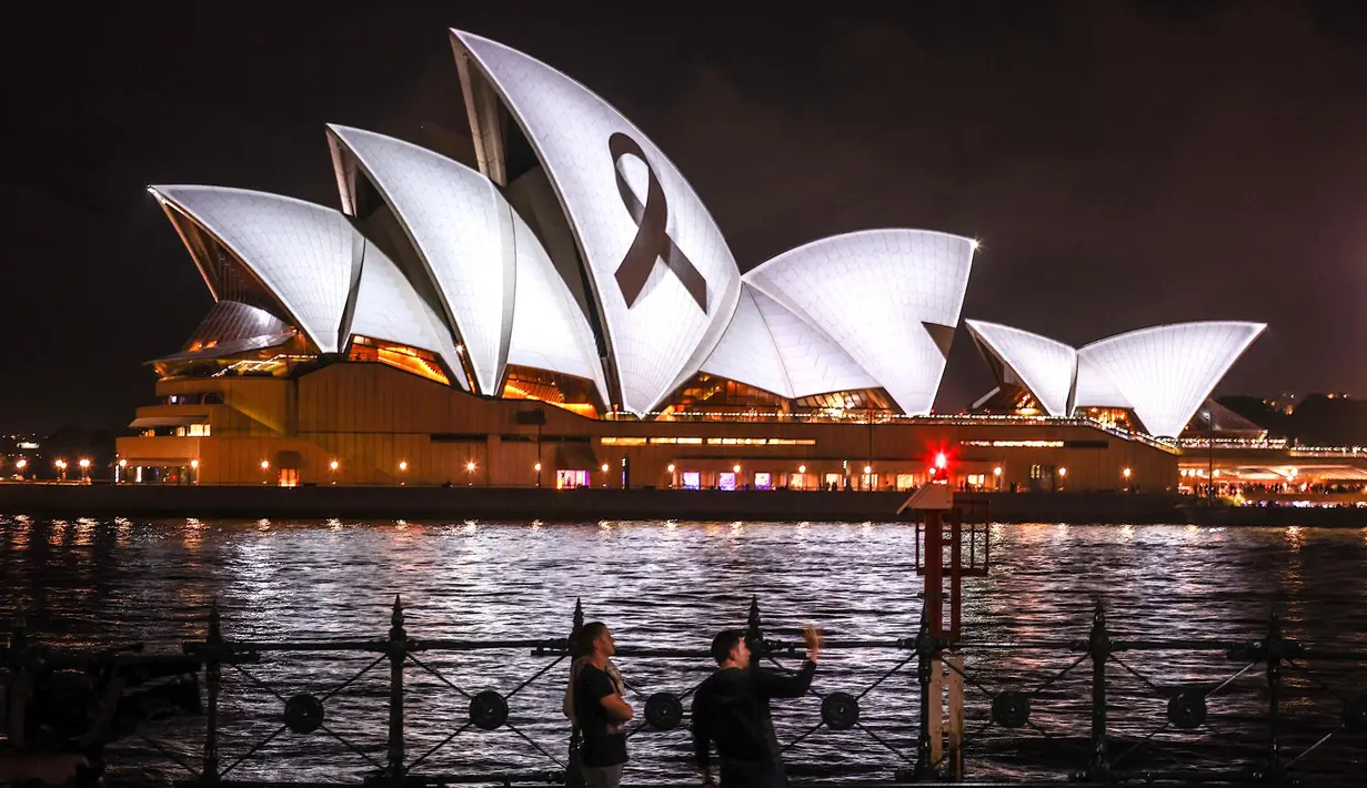 Orang-orang berdiri di depan pita hitam yang melambangkan kenangan dan duka cita, yang diproyeksikan ke Sydney Opera House, Australia, Kamis (9/2/2023). Gempa dahsyat berkekuatan 7,8 magnitudo terjadi di Turki dan Suriah pada Senin (6/2) lalu dan mengakibatkan kerusakan parah serta ribuan korban jiwa. (DAVID GRAY/AFP)