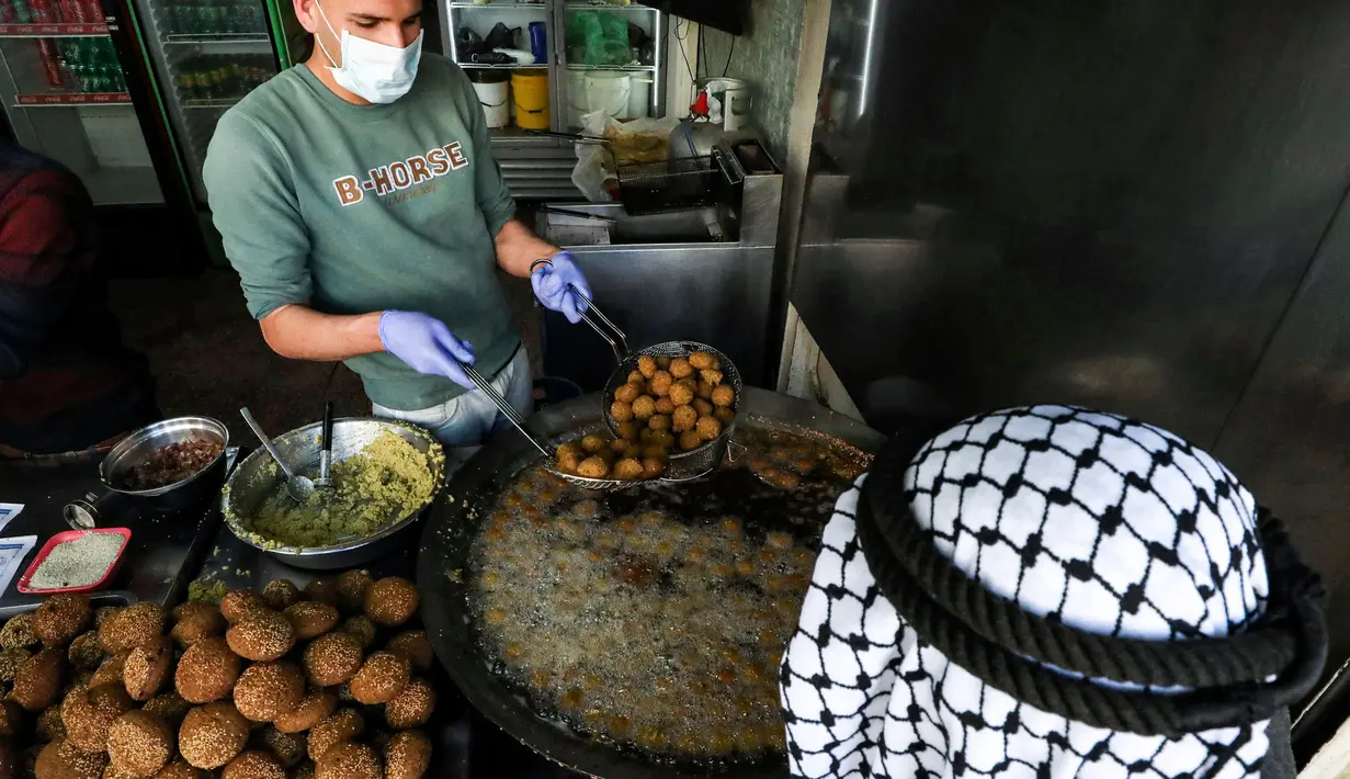 Juru masak Palestina mengenakan sarung tangan dan masker saat melayani pembeli falafel goreng di sebuah restoran lokal di kota Hebron, Tepi Barat yang diduduki pada 29 April  2020. Pada bulan suci Ramadan, falafel sering dimakan sebagai hidangan buka puasa. (HAZEM BADER / AFP)