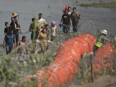 Para migran yang mencoba memasuki AS dari Meksiko mendekati lokasi di mana para pekerja sedang merakit pelampung besar yang akan digunakan sebagai penghalang perbatasan di sepanjang tepi Rio Grande di Eagle Pass, Texas, Selasa (11/7/2023). (AP Photo/Eric Gay)