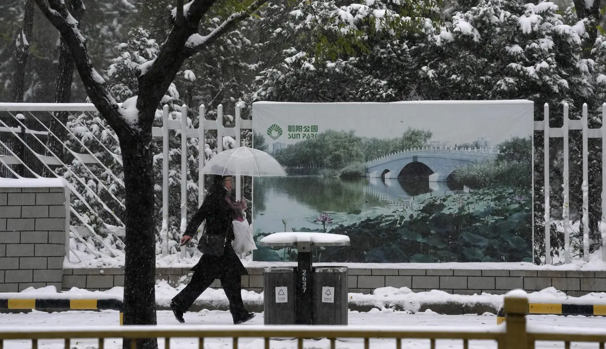 Warga berjalan melewati papan reklame taman yang menunjukkan waktu musim panas saat salju turun di Beijing, Minggu (7/11/2021). Badai salju awal musim telah menyelimuti sebagian besar China utara termasuk ibu kota Beijing, mendorong penutupan jalan dan pembatalan penerbangan. (AP Photo/Ng Han Guan)