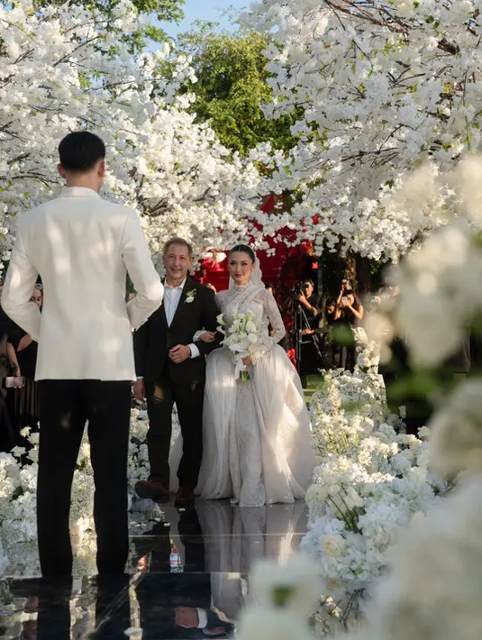 Gaun berwarna putih gading tersebut hadir dengan siluet ball gown yang megah. Bagian atasnya dirancang dengan high neckline dan lengan panjang transparan berbahan lace yang dihiasi detail payet berkilau. Permainan tekstur tulle, embroidery, dan taburan kristal menciptakan dimensi yang membuat gaun terlihat mewah dari setiap sudut. [@jennifercoppenreal20].