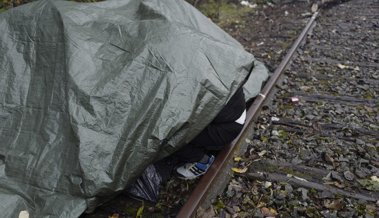 Seorang migran melindungi dirinya dari hujan di bawah terpal di kamp darurat di Calais, Prancis utara, Sabtu (277/11/2021). Di kamp-kamp darurat di luar Calais, para migran yang putus asa menunggu kesempatan untuk mencoba menyeberangi Selat Inggris. (AP Photo/Rafael Yaghobzadeh)