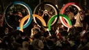 Orang-orang menunggu di dekat patung Cincin Olimpiade menjelang upacara pembukaan Olimpiade Tokyo 2020 di dekat Olympic Stadium di Tokyo, Jumat (23/7/2021). Upacara pembukaan Olimpiade Tokyo yang berlangsung dalam era pandemi digelar tanpa penonton. (Yasuyoshi CHIBA / AFP)