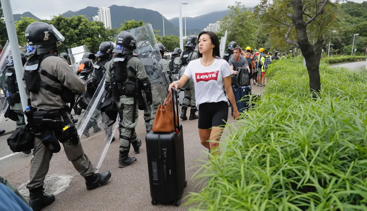 Seorang penumpang berjalan ke bandara melewati polisi anti huru-hara ketika pemrotes pro-demokrasi memblokir jalan di luar bandara di Hong Kong (1/9/2019). Demonstran pro-demokrasi penentang RUU Ekstradisi kembali menjadikan bandara sebagai tempat aksi unjuk rasa. (AP Photo/Kin Cheung)