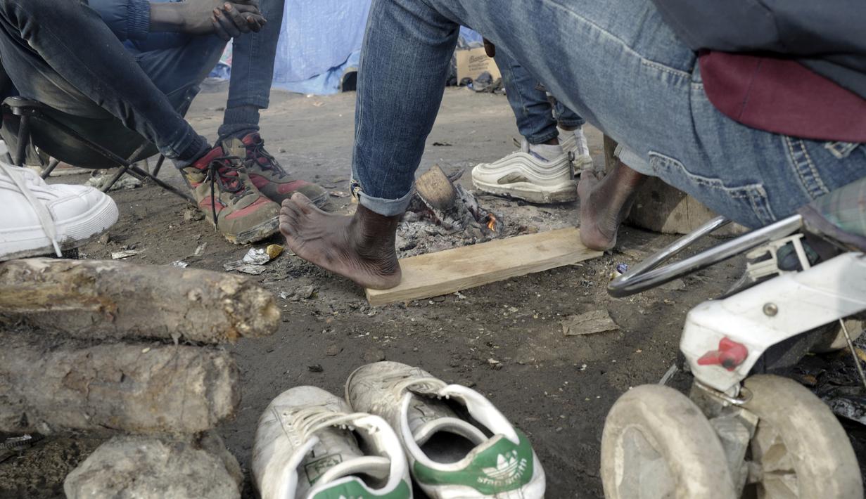 Seorang migran menghangatkan kakinya di kamp darurat di luar Calais, Prancis utara, Sabtu (277/11/2021). Di kamp-kamp darurat di luar Calais, para migran yang putus asa menunggu kesempatan untuk mencoba menyeberangi Selat Inggris. (AP Photo/Rafael Yaghobzadeh)
