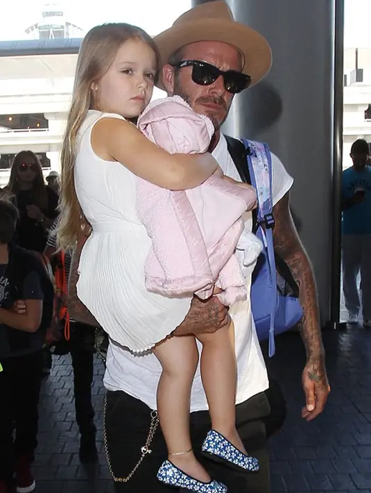 Harper Beckham digendong ayahnya saat berada di bandara LAX menuju London pada 31 Agustus 2015. Harper tampil manis dengan menggunakan dress putih polos. (harperbeckhamfashion)