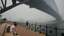 Wisatawan memakai masker saat berpose di bawah Harbour Bridge, Sydney, Australia, Selasa (10/12/2019). Kabut asap yang menyelimuti Sydney dan sebagian wilayah New South Wales dalam beberapa minggu terakhir diperkirakan akan terus berlangsung selama beberapa bulan ke depan. (TAMAN PETER/AFP)