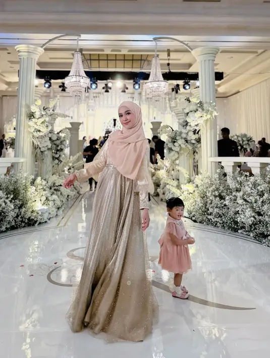 Potret kondangan menggemaskan dari Ria Ricis dan Moana. Ricis tampil cantik penuh pesona dengan dress emas yang serasi dengan hijabnya. Sedangkan Moana mengenakan dress cokelat yang tak kalah manis. [Foto: Instagram/riaricis1795]