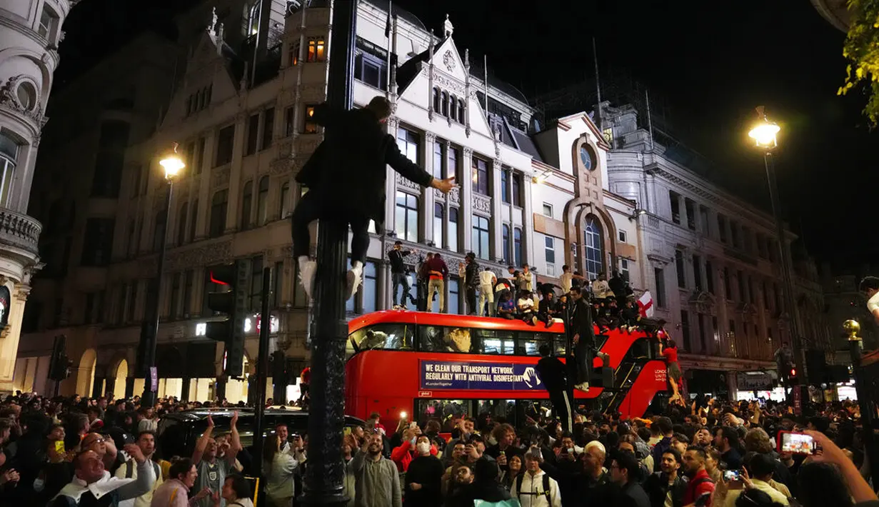 Seorang pria memanjat tiang lampu saat penggemar merayakan di atas bus di pusat kota London setelah Inggris memenangkan semifinal Euro 2020 antara Inggris dan Denmark yang dimainkan di Stadion Wembley (8/7/2021). Inggris di laga final berhadapan dengan Italia. (AP Photo/Thanassis Stavrakis)