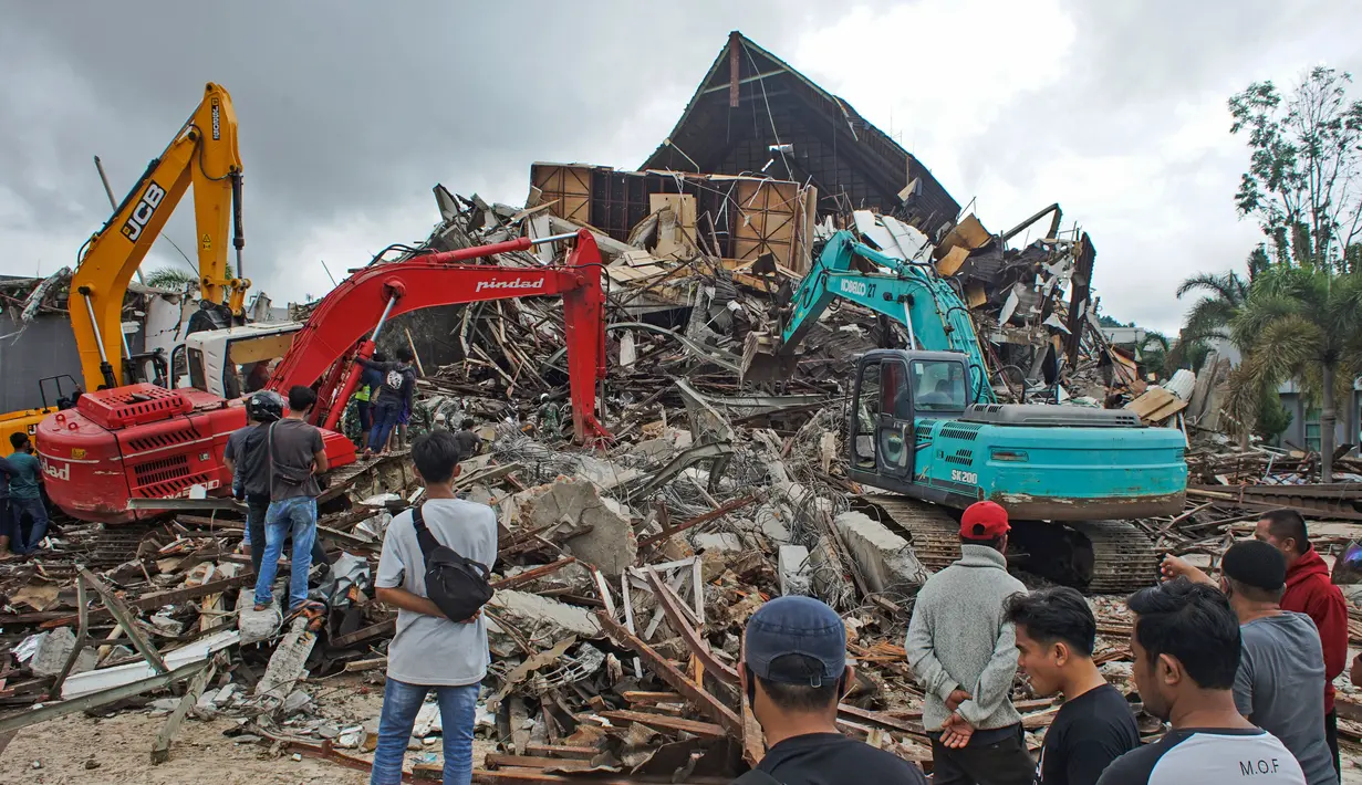 Sejumlah warga menyaksikan alat berat mencari korban di reruntuhan gedung pemerintah daerah yang runtuh saat gempa bumi di Mamuju, Sulawesi Barat, Jumat (15/1/2021). (AP Photo/Azhari Surahman)