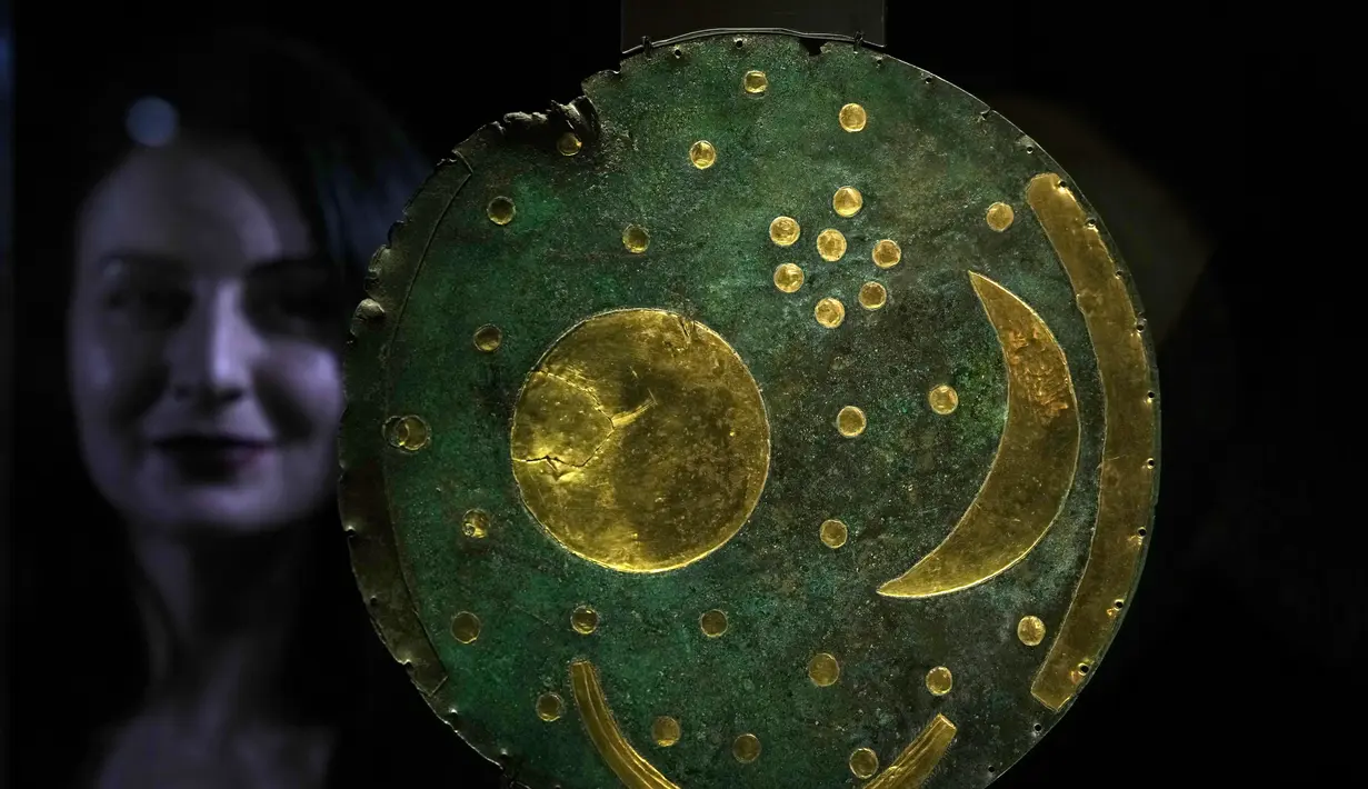 Seorang anggota staf berpose di sebelah 'Nebra Sky Disc' yang berasal dari sekitar tahun 1600 SM, dan merupakan representasi kosmos tertua yang masih ada, dipamerkan di pameran The World of Stonehenge' di British Museum di London, Senin (14/2/2022). (AP Photo/Alastair Grant)