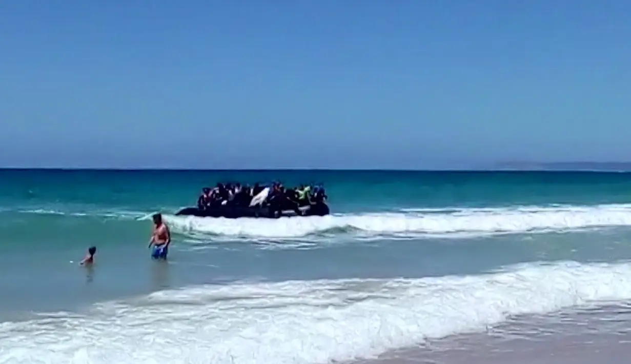 Seorang pria dan anak laki-laki menyaksikan  perahu karet yang dipenuhi imigran mendekati tepi pantai Cadiz di Spanyol selatan, 9 Agustus 2017. Puluhan imigran tersebut langsung melompat ke air dan berlarian menuju pantai. (Carlos Sanz via AP)