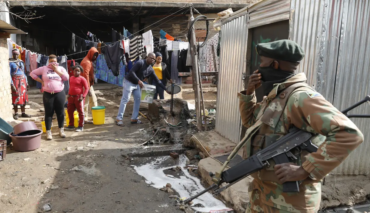 Orang-orang berdiri dekat rumah mereka dan menyaksikan anggota Pasukan Pertahanan Afrika Selatan (SANDF) berpatroli di daerah mereka di Alexandra, Johannesburg, Kamis (15/7/2021). Patroli tersebut menyusul kerusuhan massa yang disebabkan dipenjaranya mantan Presiden Jacob Zuma. (Phill Magakoe/AFP)