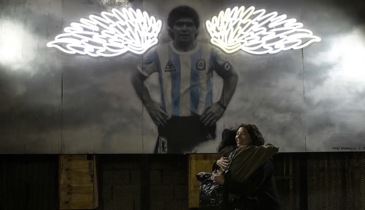 Perempuan berpelukan di dekat mural mendiang legenda sepak bola dunia, Diego Maradona di Buenos Aires, Argentina, Selasa malam (23/11/2021). Peringatan pertama kematian Maradona digelar pada 25 November 2021. (AP Photo/Rodrigo Abd)