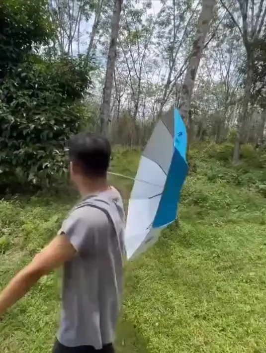 Tak berhenti sampai di situ, Jirayut terlihat berdiri santai sambil memegang payung di bawah terik matahari. Latar belakangnya tetap kebun karet yang rindang dan hijau, menambah kesan sejuk di tengah cuaca panas. Ia menceritakan bahwa luas kebun karetnya hampir mencapai satu hektar.(Liputan6.com/IG/@jirayutdaa4official)
