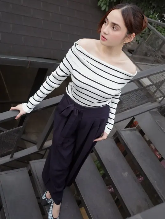 Syifa mengenakan atasan off shoulder warna putih bergaris hitam dipadukan celana hitam panjang belt tali. Ia tampil dengan flat shoes berpita dan anting-anting emas. [@syifahadju]