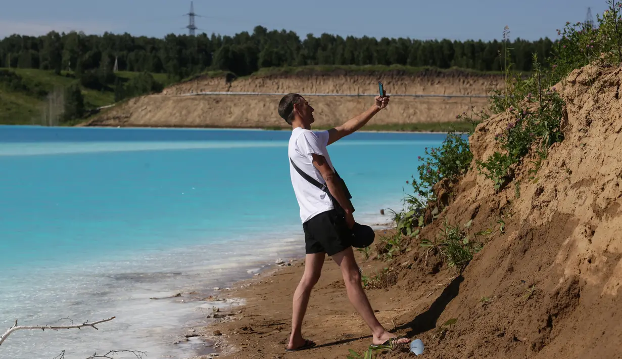 Pengunjung berswafoto dengan latar belakang tempat pembuangan abu dari pabrik batubara lokal di Danau Novosibirsk, Rusia, Kamis (11/7/2019). Dasar danau itu cukup dalam dan berlumpur, sehingga jika sudah tenggelam akan sulit menyelamatkan diri. (Rostislav NETISOV/AFP)